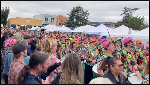 Photo of a parade with drummers in florescent  pinks, greens and oranges, marching down a street in front of tents.