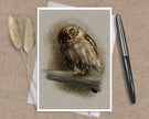 A square art print featuring a detailed illustration of a brown owl perched on a branch, displayed on a textured brown surface with a black pen and dried grass stems as props.