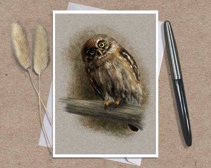 A square art print featuring a detailed illustration of a brown owl perched on a branch, displayed on a textured brown surface with a black pen and dried grass stems as props.