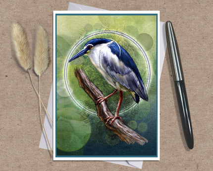A square art print featuring a detailed illustration of a blue and white heron perched on a branch against a green bokeh background, styled on a brown textured surface with a black pen and dried grass stems.