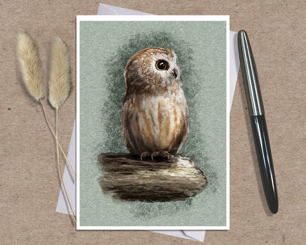A square art print featuring a detailed illustration of a brown and beige owl perched on a log, set against a muted sage green background. The print is styled in a flat-lay on a brown textured surface, accompanied by a black pen on the right and dried bunny tail grass on the left.