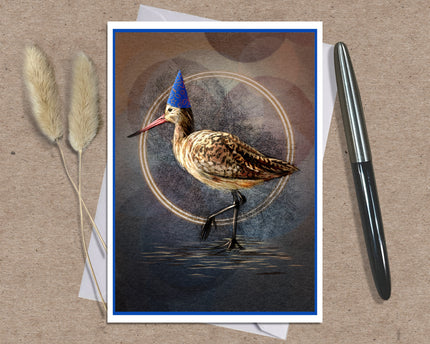Product photo of a greeting card featuring a Marbled Godwit in a blue conical party hat. Beside the card are two atmosphere props--a spring of grasses and an ink pen.