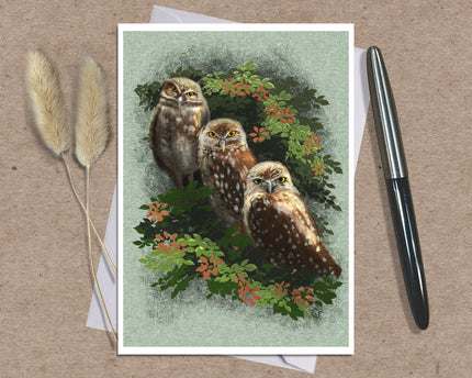 A square art print featuring three owls among green leaves and small orange flowers, styled on a kraft paper background with a white envelope, dried grass stems, and a black pen.