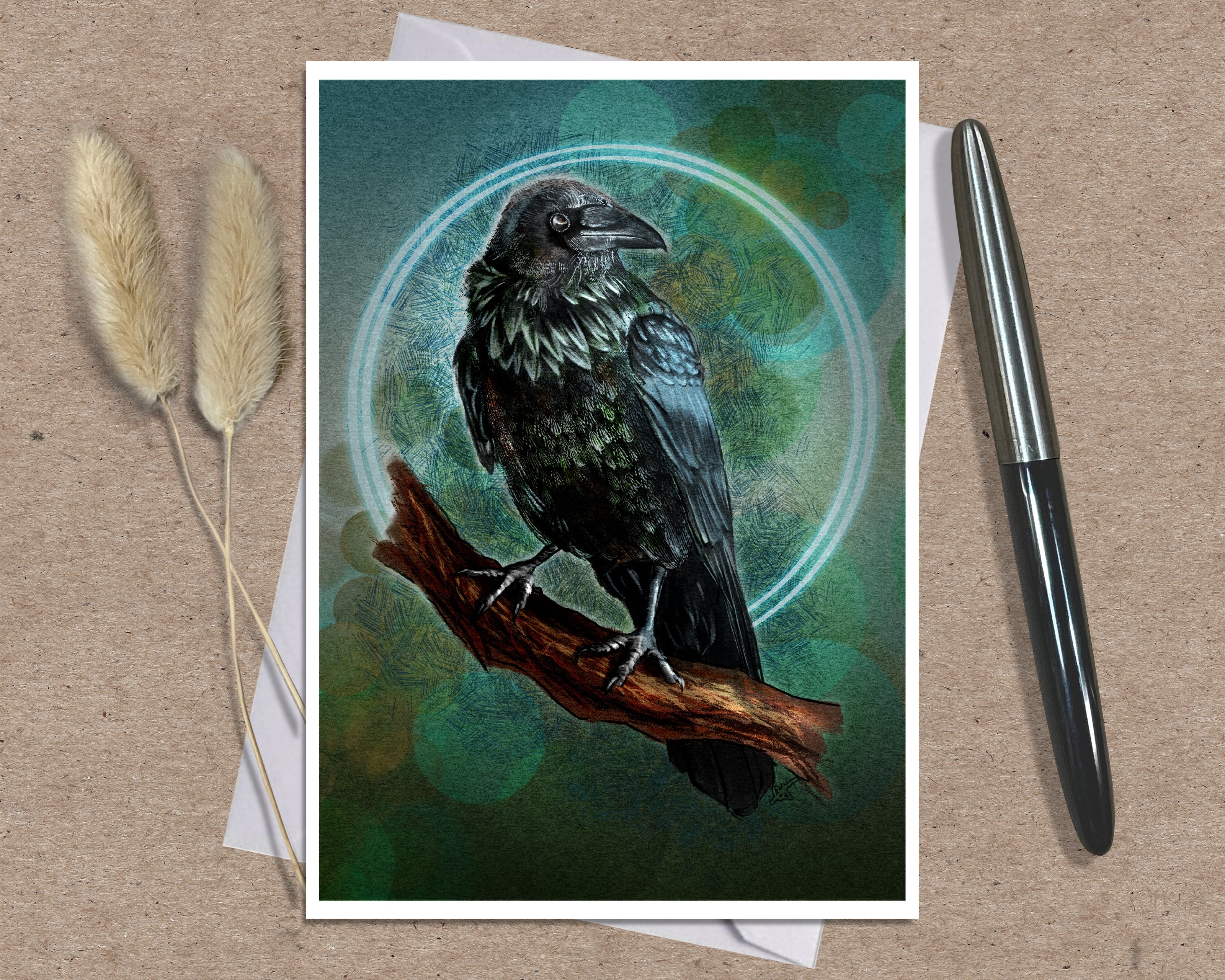 A greeting card art print featuring a detailed illustration of a black raven perched on a brown branch against a teal and green circular background, styled on a kraft paper surface with a silver pen and dried grass stems.