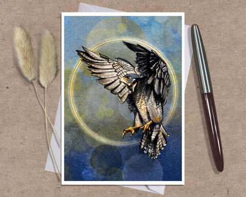 A greeting card featuring a detailed illustration of a hawk with wings spread, set against a blue watercolor background with a gold circular halo. The print is displayed on a textured brown surface next to a silver and brown pen and two dried beige grasses.