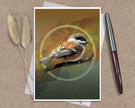 An art print greeting card featuring a detailed bird illustration perched on a branch, displayed on a kraft paper background with two dried grass stems on the left and a silver pen on the right.