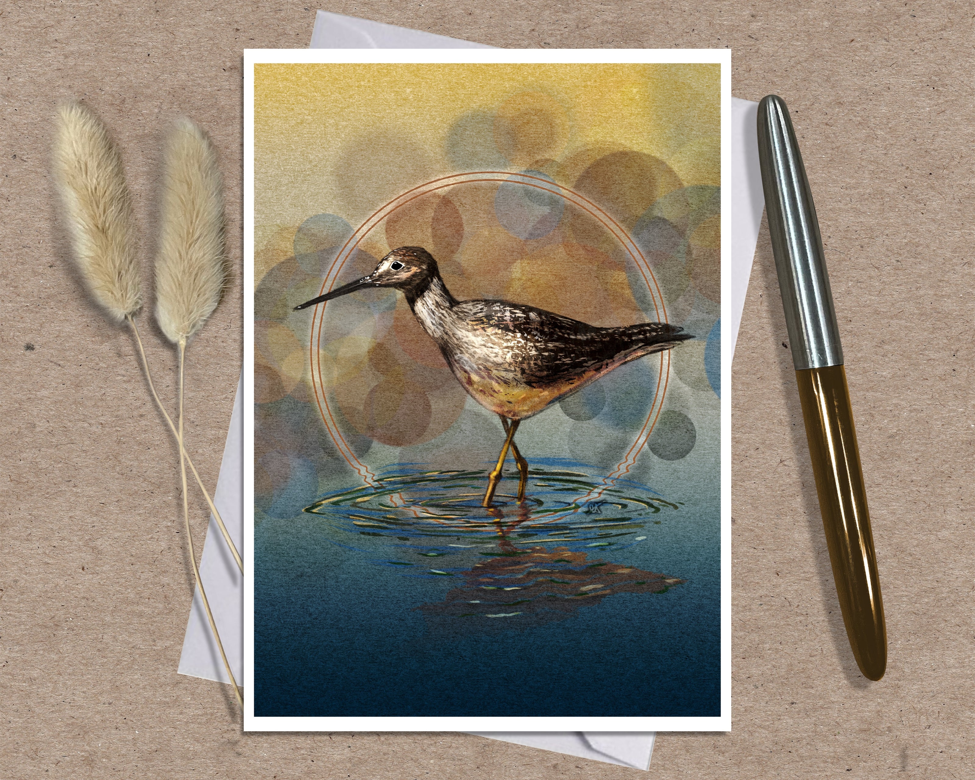 An art print greeting card featuring a detailed illustration of a wading bird standing in water, styled on a textured brown surface with a silver and gold pen and dried grass stems.