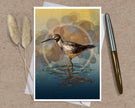 An art print greeting card featuring a detailed illustration of a wading bird standing in water, styled on a textured brown surface with a silver and gold pen and dried grass stems.