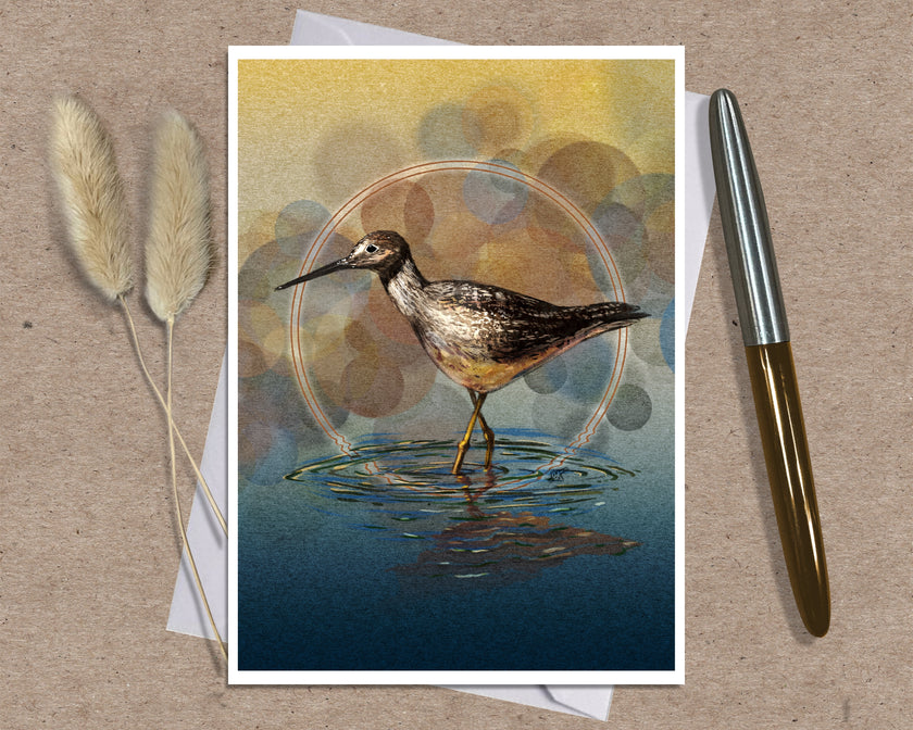 An art print greeting card featuring a detailed illustration of a wading bird standing in water, styled on a textured brown surface with a silver and gold pen and dried grass stems.