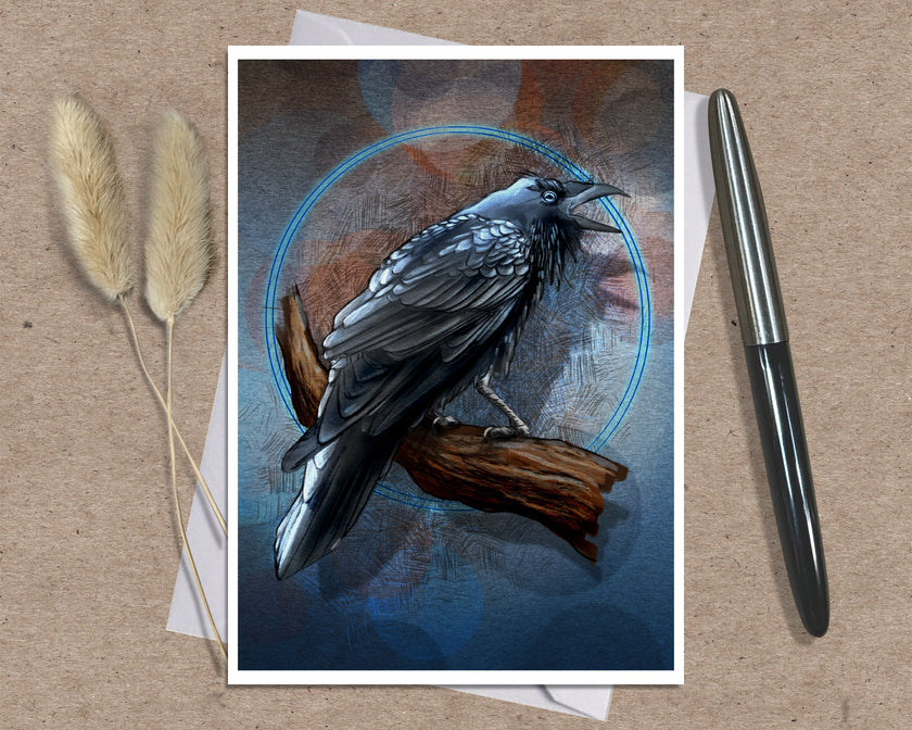 An art print greeting card featuring a detailed illustration of a black raven perched on a branch against a dark blue and brown background with a glowing blue circle, styled on a kraft paper surface with dried grass stems and a silver pen.