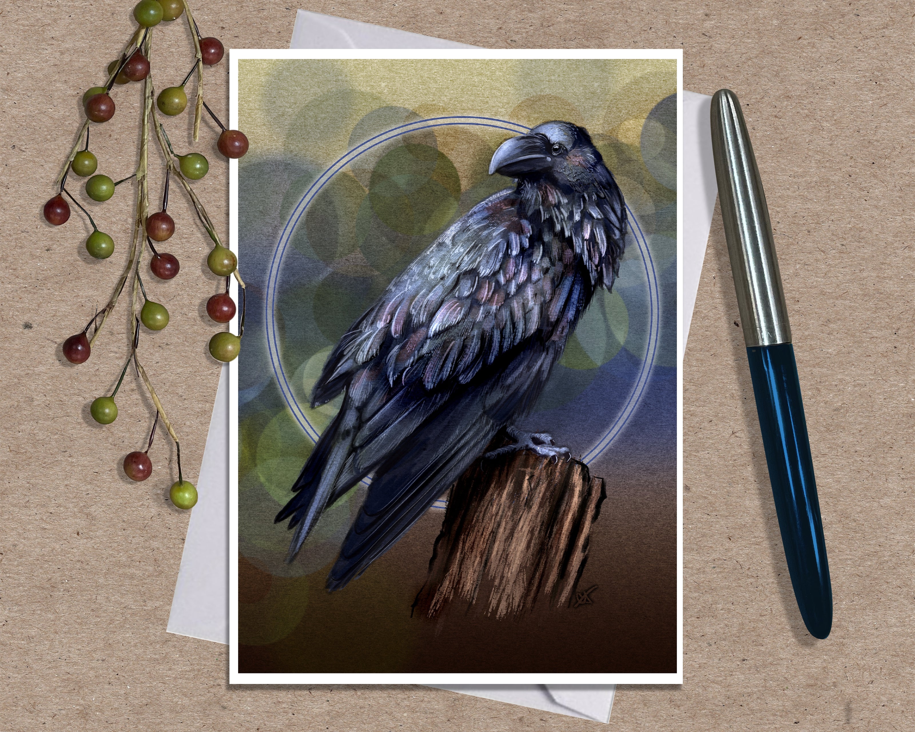 An art print greeting card featuring a detailed illustration of a black raven perched on a branch against a yellow, blue and brown background with a glowing blue circle, styled on a kraft paper surface with dried grass stems and a silver pen.