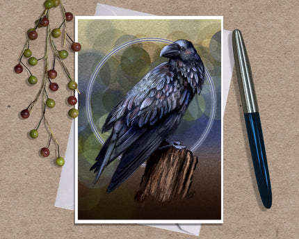 An art print greeting card featuring a detailed illustration of a black raven perched on a branch against a yellow, blue and brown background with a glowing blue circle, styled on a kraft paper surface with dried grass stems and a silver pen.