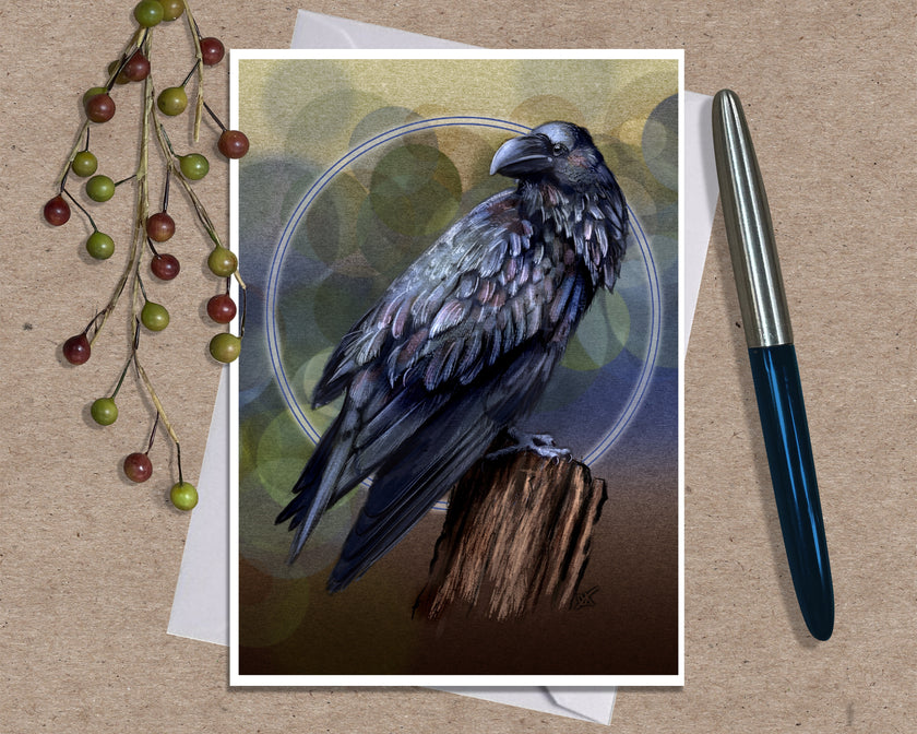 An art print greeting card featuring a detailed illustration of a black raven perched on a branch against a yellow, blue and brown background with a glowing blue circle, styled on a kraft paper surface with dried grass stems and a silver pen.