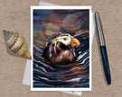 An art print greeting card featuring a painted puffin in dark blue water, styled on a brown kraft paper background with a seashell on the left and a silver pen on the right.