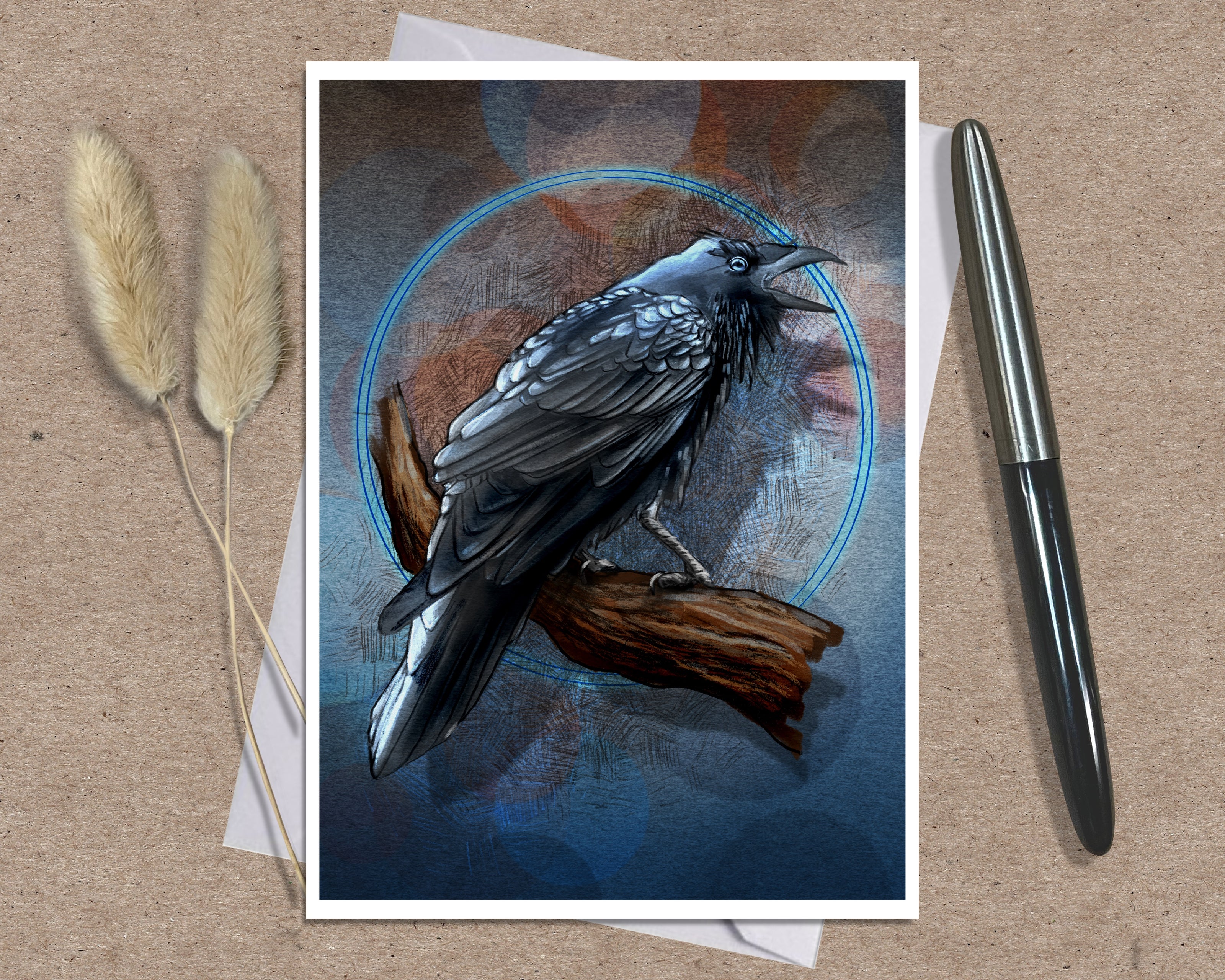 An art print greeting card featuring a detailed illustration of a black raven perched on a branch against a dark blue and brown background with a glowing blue circle, styled on a kraft paper surface with dried grass stems and a silver pen.