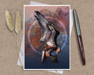 An art print greeting card featuring a detailed illustration of a bird of prey with wings spread, set against an abstract textured background in blue, brown, and orange tones. The print is displayed on a kraft paper background alongside two dried grass stems and a silver and brown pen.