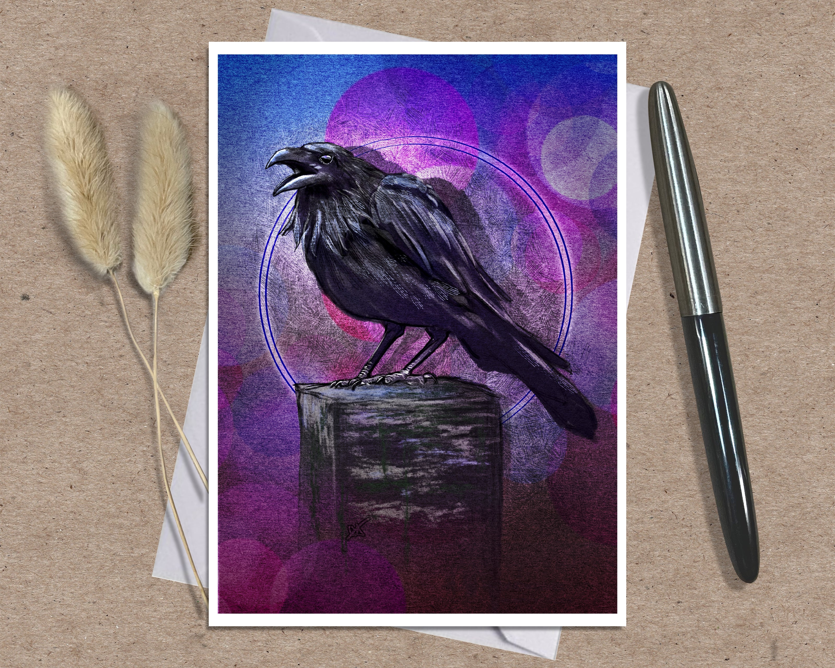 An art greeting card print featuring a black raven perched on a wooden block against a purple and blue abstract background, styled on a kraft paper surface with dried grass stems and a pen.