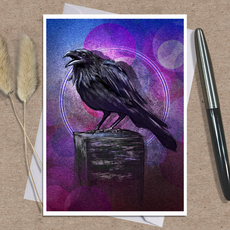 An art greeting card print featuring a black raven perched on a wooden block against a purple and blue abstract background, styled on a kraft paper surface with dried grass stems and a pen.