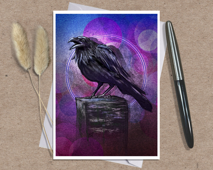 An art greeting card print featuring a black raven perched on a wooden block against a purple and blue abstract background, styled on a kraft paper surface with dried grass stems and a pen.
