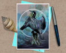 An art print greeting card featuring a dark bird perched on a rock against a blue-green bokeh background with a white circular frame, styled on a kraft paper surface with a turquoise card, a seashell, and a blue pen.