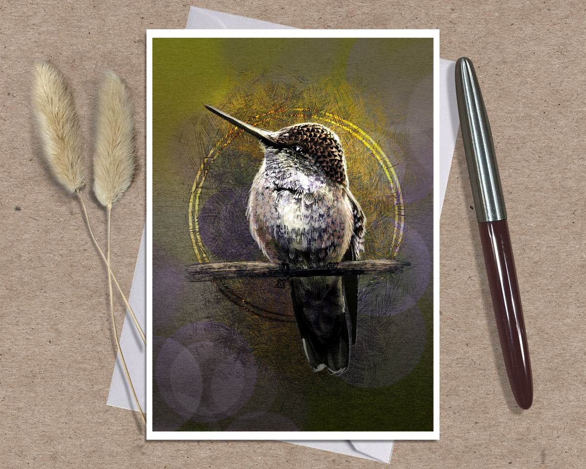 A greeting card featuring a detailed hummingbird perched on a branch against an abstract olive-green and gold background, styled on a kraft paper surface with a white pen and dried bunny tail grass.