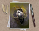 A greeting card featuring a detailed hummingbird perched on a branch against an abstract olive-green and gold background, styled on a kraft paper surface with a white pen and dried bunny tail grass.