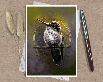 A greeting card featuring a detailed hummingbird perched on a branch against an abstract olive-green and gold background, styled on a kraft paper surface with a white pen and dried bunny tail grass.