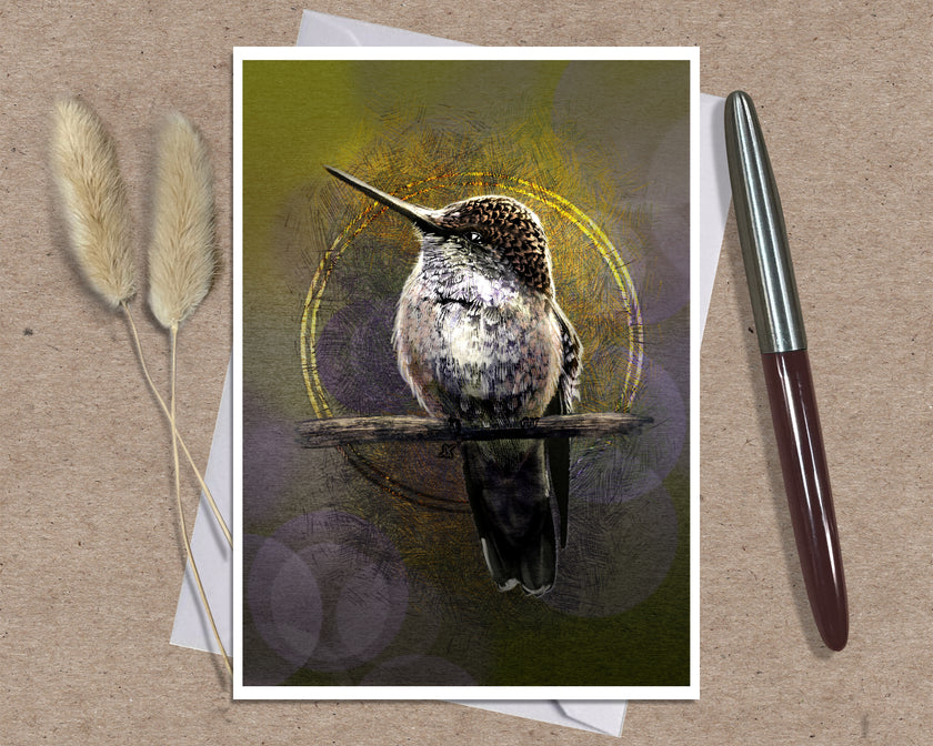 A greeting card featuring a detailed hummingbird perched on a branch against an abstract olive-green and gold background, styled on a kraft paper surface with a white pen and dried bunny tail grass.