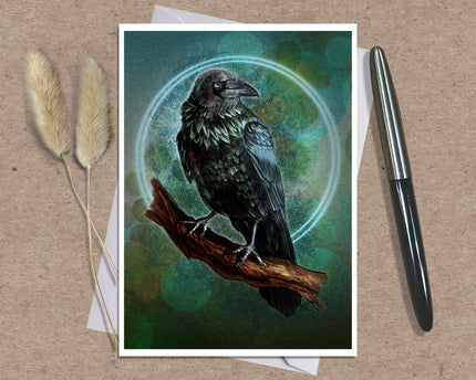 A greeting card art print featuring a detailed illustration of a black raven perched on a brown branch against a teal and green circular background, styled on a kraft paper surface with a silver pen and dried grass stems.