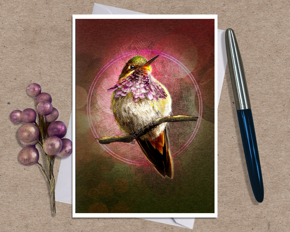 A art print greeting card featuring a hummingbird perched on branches with a glowing circular motif and pink-toned background, styled on a kraft paper surface with a white envelope and a black pen nearby.