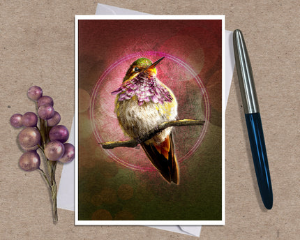 A art print greeting card featuring a hummingbird perched on branches with a glowing circular motif and pink-toned background, styled on a kraft paper surface with a white envelope and a black pen nearby.