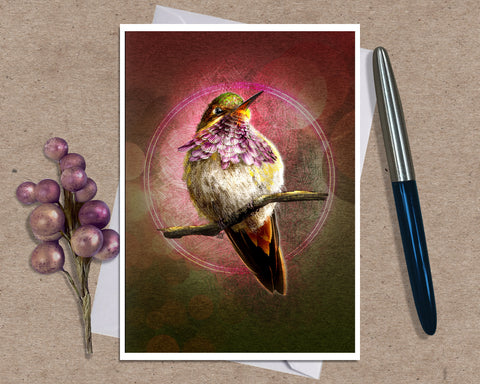 A art print greeting card featuring a hummingbird perched on branches with a glowing circular motif and pink-toned background, styled on a kraft paper surface with a white envelope and a black pen nearby.