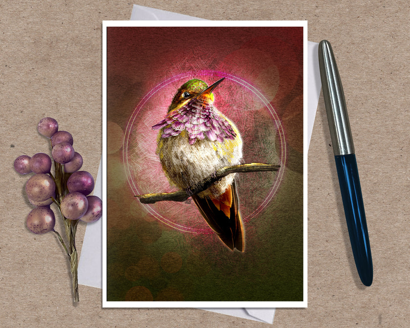 A art print greeting card featuring a hummingbird perched on branches with a glowing circular motif and pink-toned background, styled on a kraft paper surface with a white envelope and a black pen nearby.