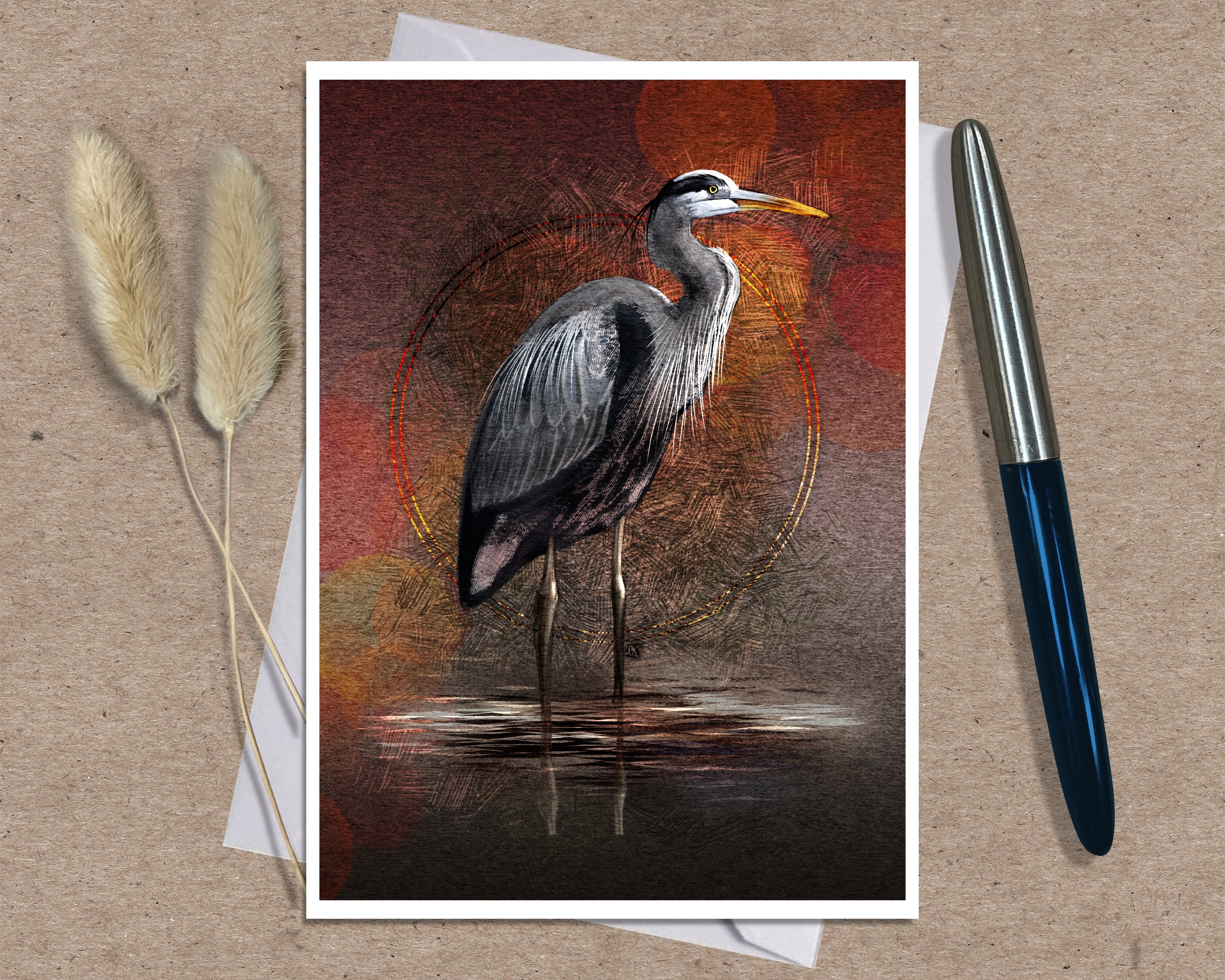 An art print greeting card featuring a heron standing in shallow water against a warm red, orange, and brown textured background, styled on a kraft paper surface with dried grass stems and a pen nearby.