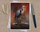 An art print greeting card featuring a heron standing in shallow water against a warm red, orange, and brown textured background, styled on a kraft paper surface with dried grass stems and a pen nearby.