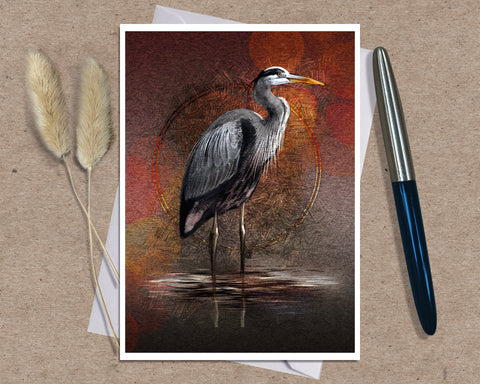 An art print greeting card featuring a heron standing in shallow water against a warm red, orange, and brown textured background, styled on a kraft paper surface with dried grass stems and a pen nearby.