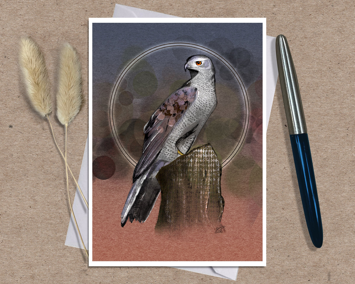 An art print greeting card featuring a detailed illustration of a bird of prey perched on a tree stump, set against a textured background with abstract circular patterns. The print is styled on a brown kraft paper surface with a white envelope underneath, flanked by two dried bunny tails on the left and a silver and blue pen on the right.