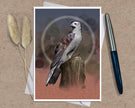 An art print greeting card featuring a detailed illustration of a bird of prey perched on a tree stump, set against a textured background with abstract circular patterns. The print is styled on a brown kraft paper surface with a white envelope underneath, flanked by two dried bunny tails on the left and a silver and blue pen on the right.