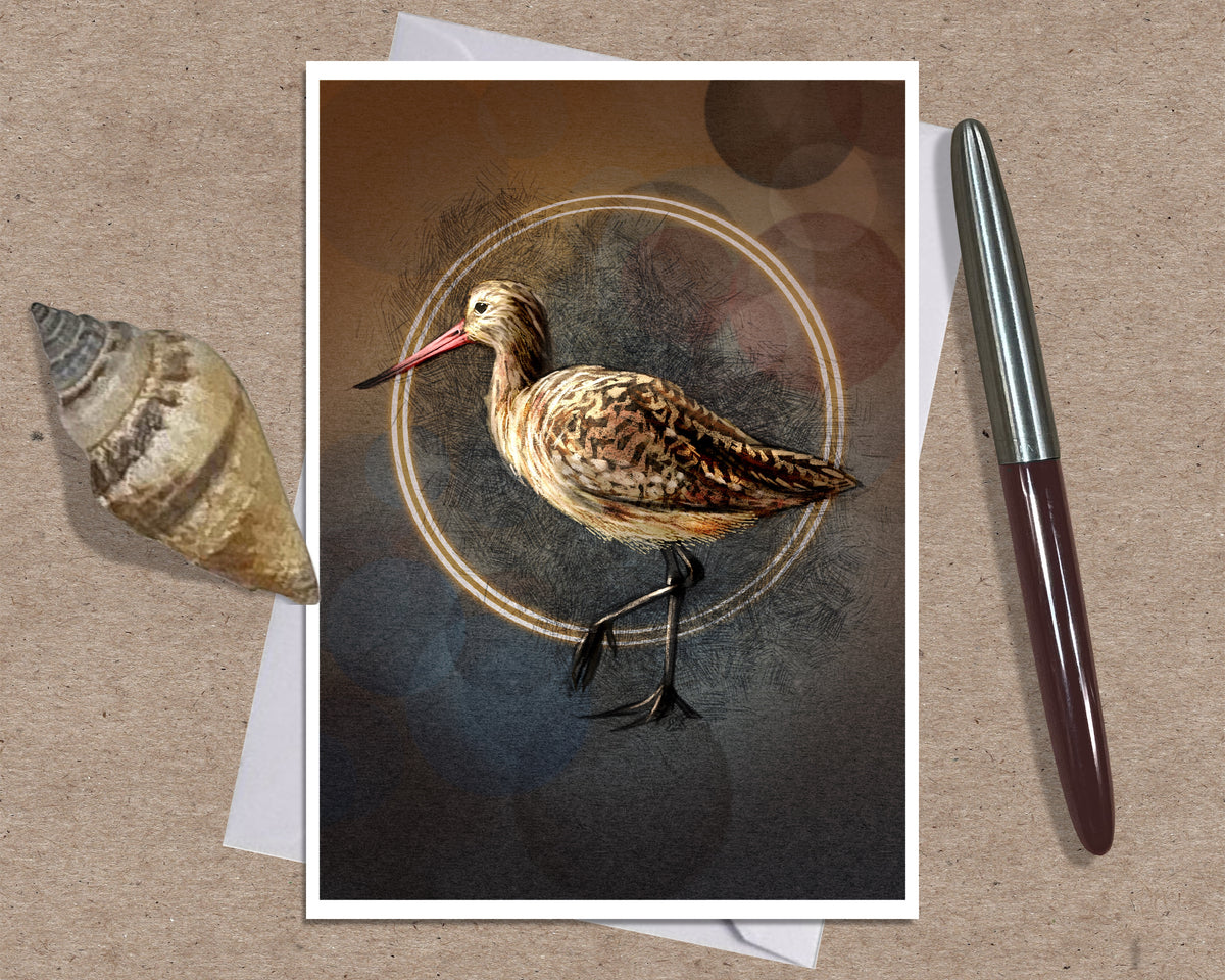 A greeting card featuring a detailed illustration of a brown and beige shorebird with a long pink beak, standing against a dark textured background with a circular gold ring. The print is displayed on a kraft paper background alongside a seashell and a silver pen.