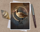 A greeting card featuring a detailed illustration of a brown and beige shorebird with a long pink beak, standing against a dark textured background with a circular gold ring. The print is displayed on a kraft paper background alongside a seashell and a silver pen.