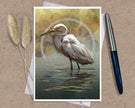An art print greeting card featuring a white egret standing in shallow water with a circular halo motif, styled on a brown textured surface with a silver pen and dried grass stems.