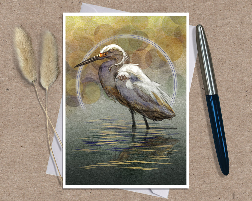 An art print greeting card featuring a white egret standing in shallow water with a circular halo motif, styled on a brown textured surface with a silver pen and dried grass stems.