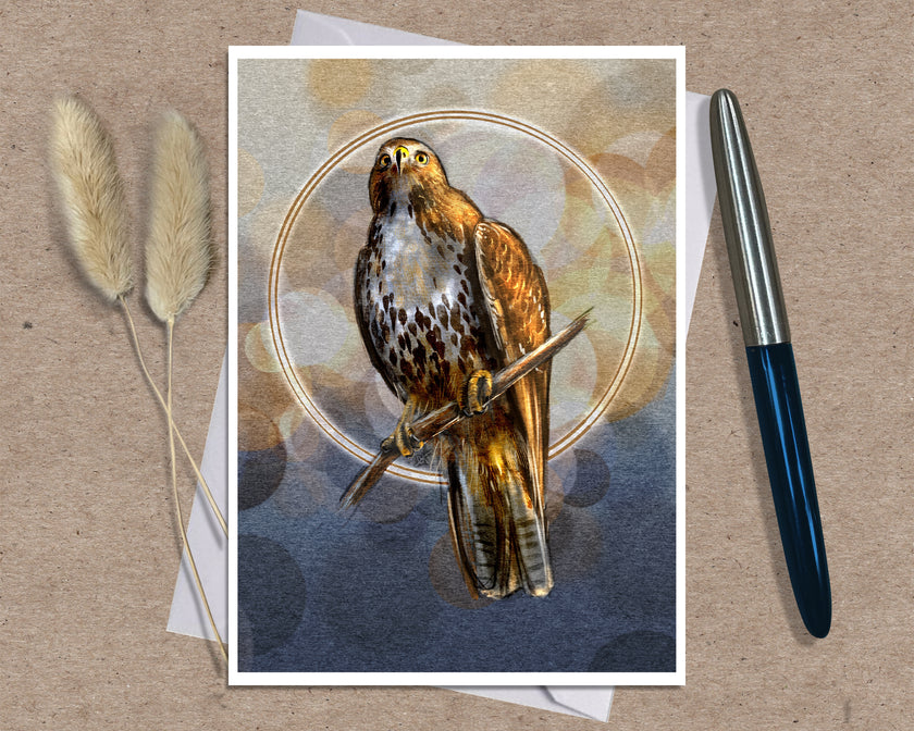 An art print greeting card featuring a detailed illustration of a hawk perched on a branch, set against a circular gold halo and abstract background. The print is styled on a brown kraft paper background with a white envelope underneath, flanked by two dried bunny tails on the left and a silver and blue pen on the right.