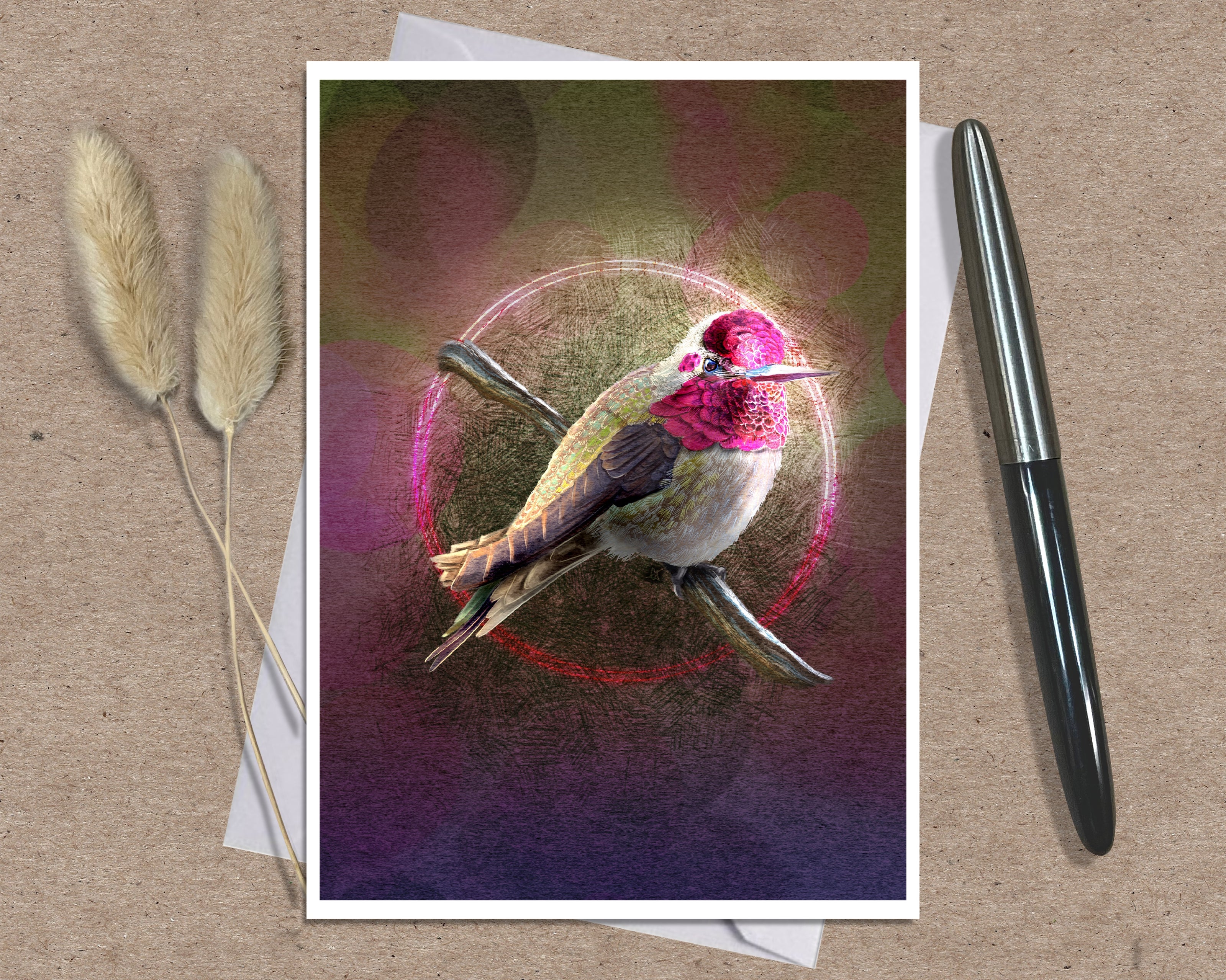 A art print greeting card featuring a hummingbird perched on branches with a glowing circular motif and pink-toned background, styled on a kraft paper surface with a white envelope and a black pen nearby.