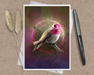 A art print greeting card featuring a hummingbird perched on branches with a glowing circular motif and pink-toned background, styled on a kraft paper surface with a white envelope and a black pen nearby.