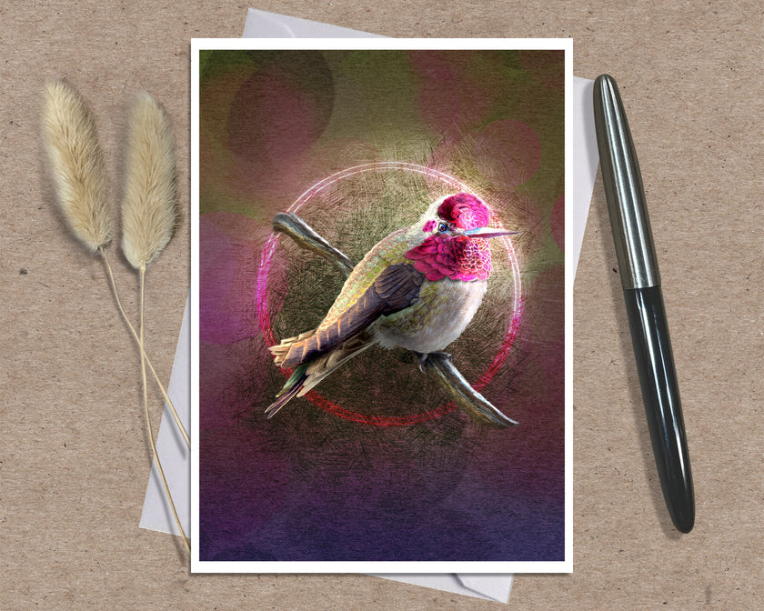 A art print greeting card featuring a hummingbird perched on branches with a glowing circular motif and pink-toned background, styled on a kraft paper surface with a white envelope and a black pen nearby.