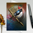 Card with a bird wearing a party hat on a branch, surrounded by colorful abstract designs.