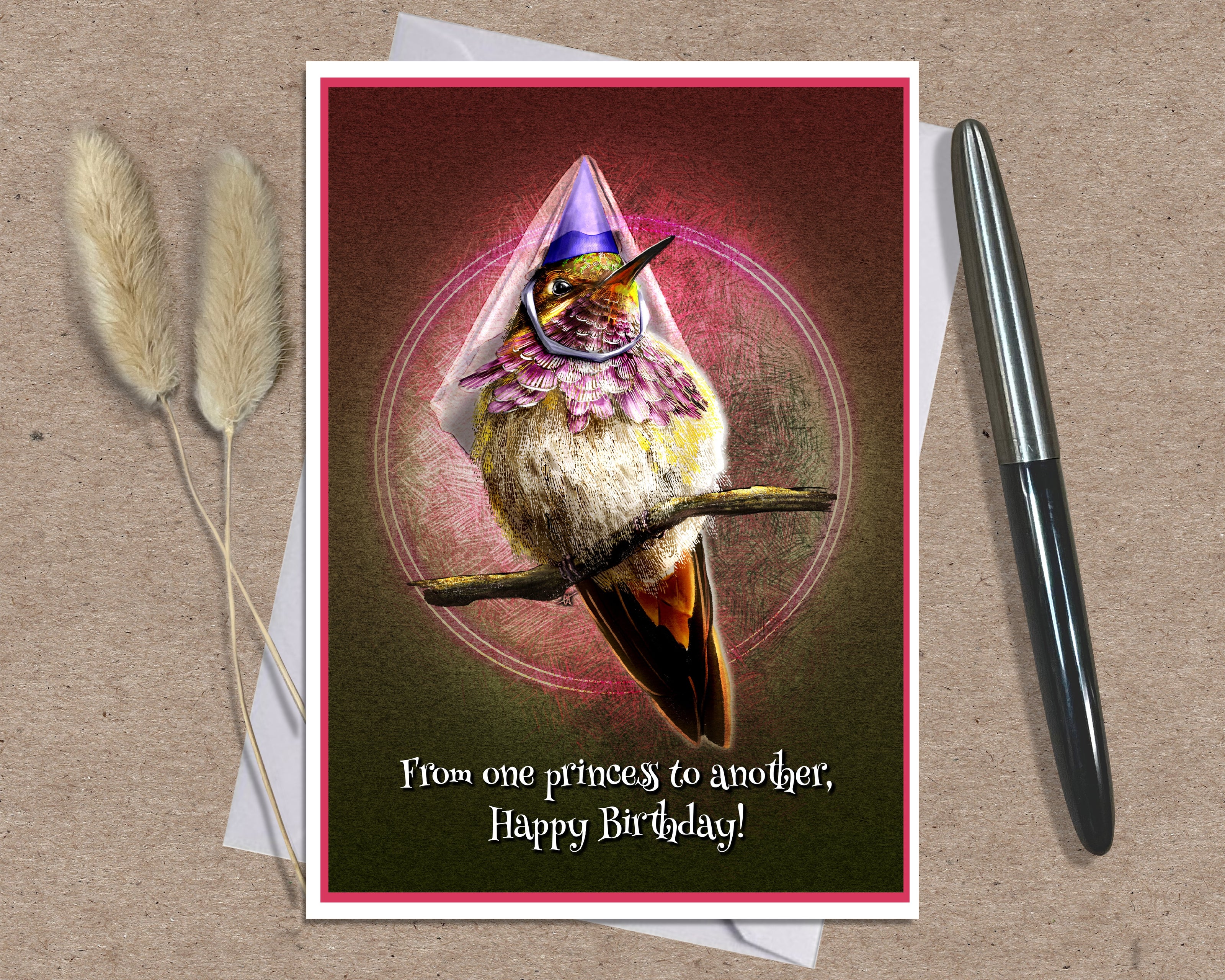 A birthday card featuring a whimsical hummingbird wearing a purple party hat and pink bow, displayed on a kraft paper background with a white envelope, two dried bunny tails, and a black pen.