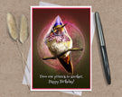 A birthday card featuring a whimsical hummingbird wearing a purple party hat and pink bow, displayed on a kraft paper background with a white envelope, two dried bunny tails, and a black pen.