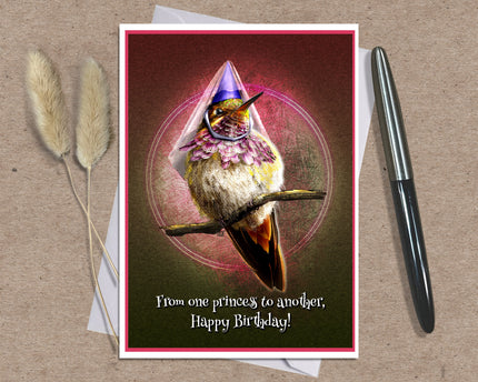 A birthday card featuring a whimsical hummingbird wearing a purple party hat and pink bow, displayed on a kraft paper background with a white envelope, two dried bunny tails, and a black pen.